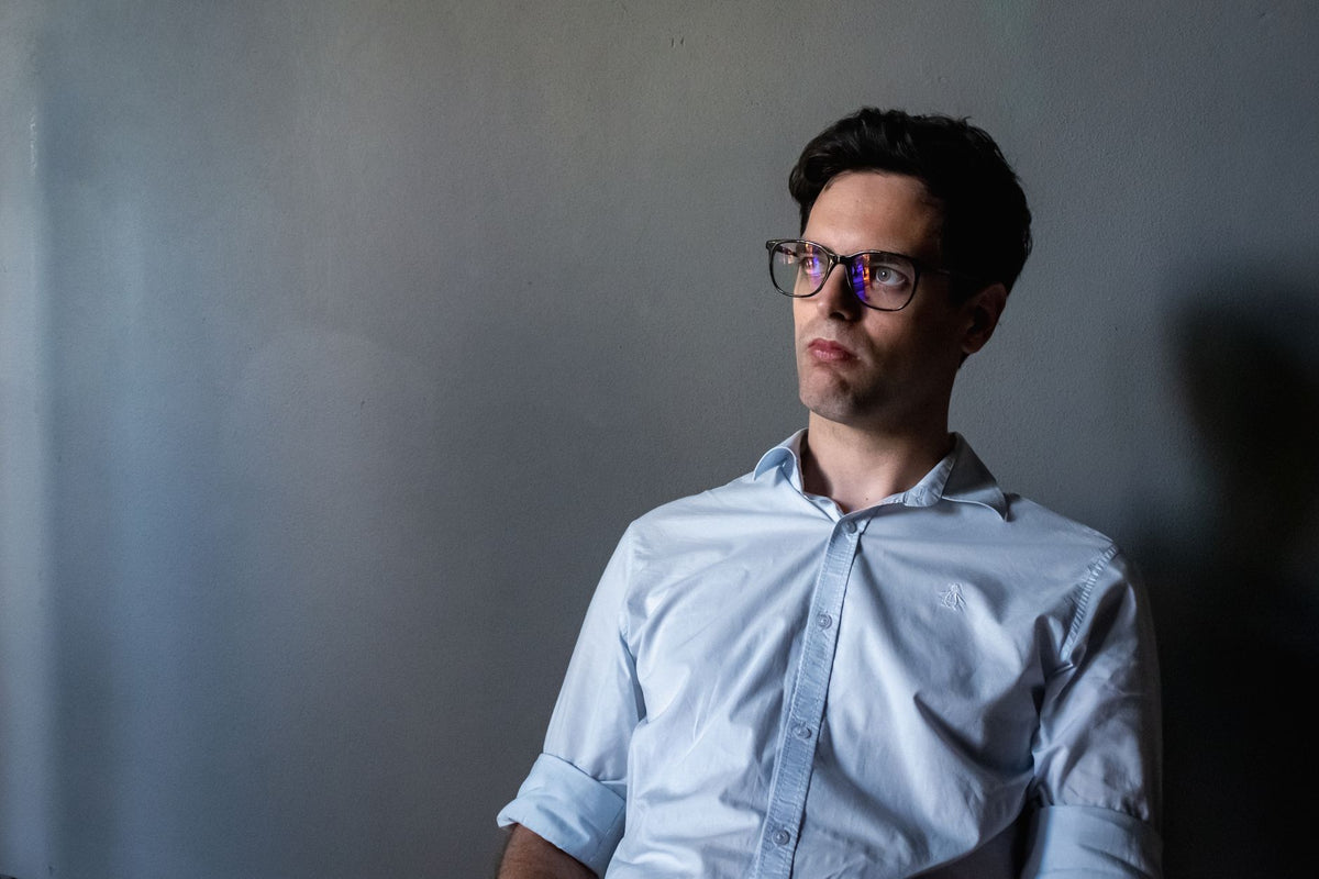 A thoughtful man with glasses sits in a dimly lit room, wearing a light blue shirt and gazing intently.