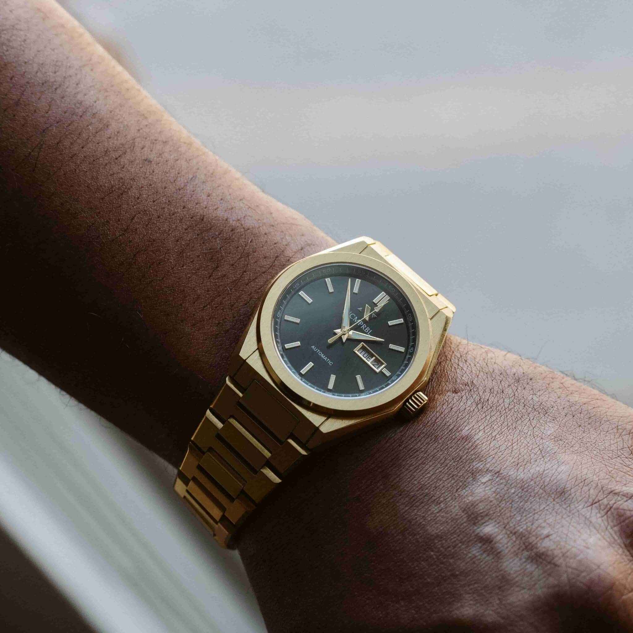 A gold-tone wristwatch with a dark blue dial and date window, worn on a person's wrist against a soft gray background.