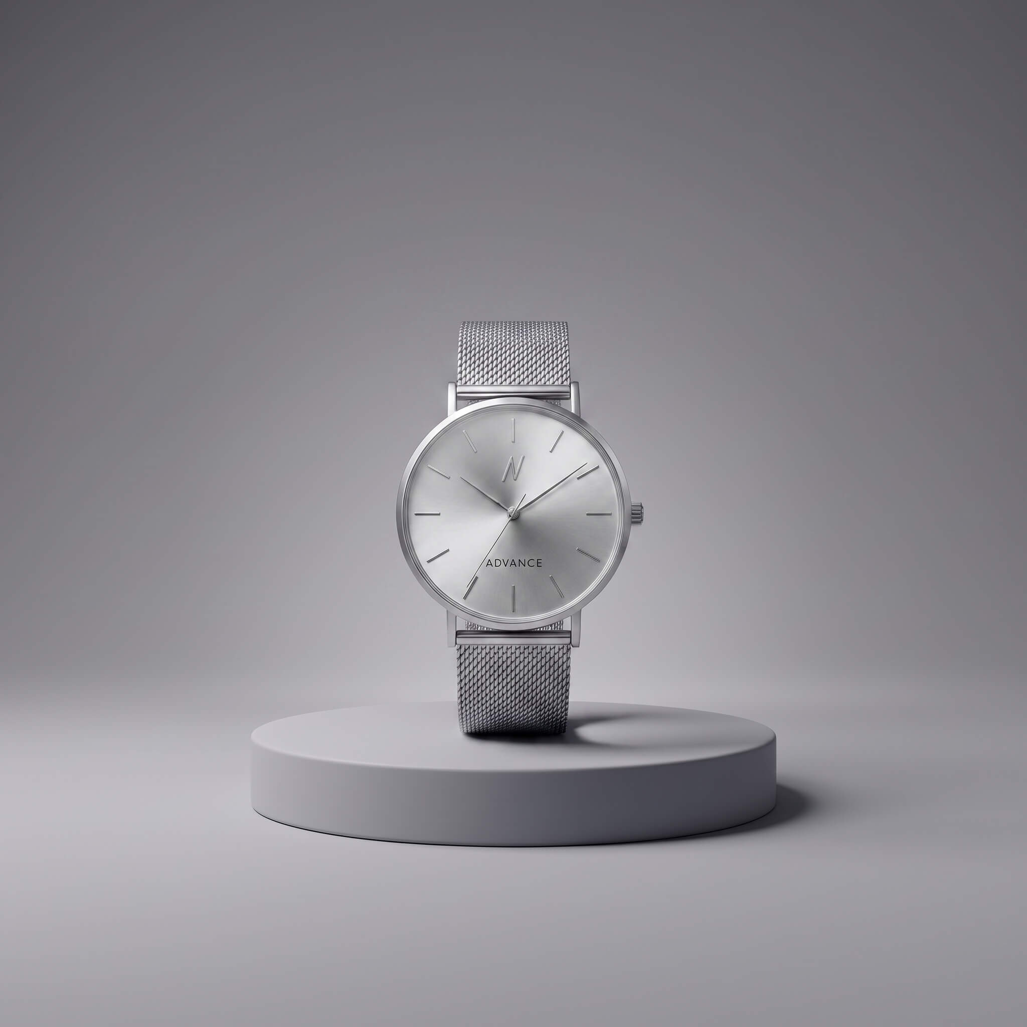 A minimalist silver analog watch with a mesh strap and a silver dial, displayed on a round gray pedestal against a clean gray background. The brand name 'ADVANCE' is visible on the dial.
