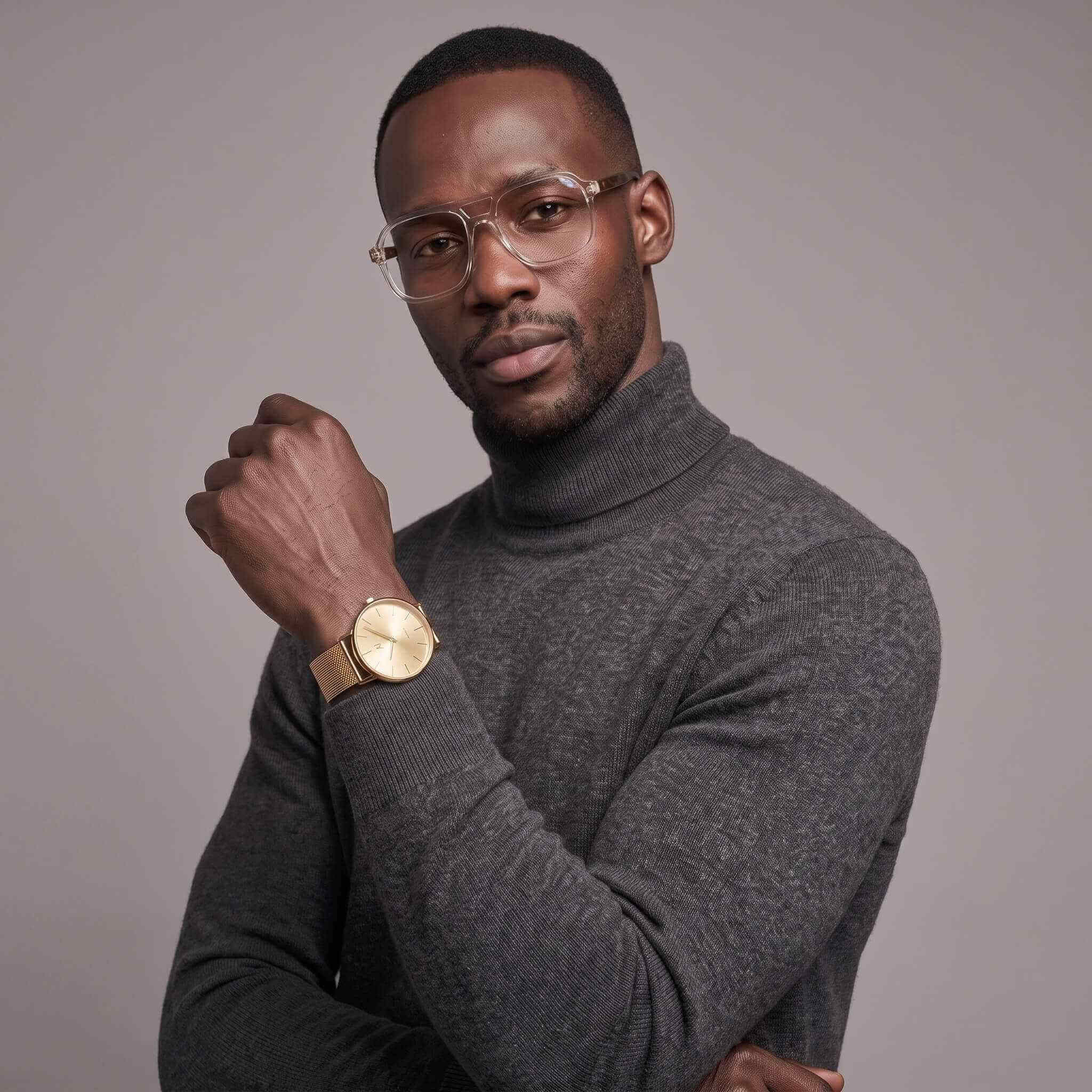 A man wearing a dark gray turtleneck sweater and clear-framed eyeglasses, with a gold-tone watch on his wrist, posed against a solid gray studio background.