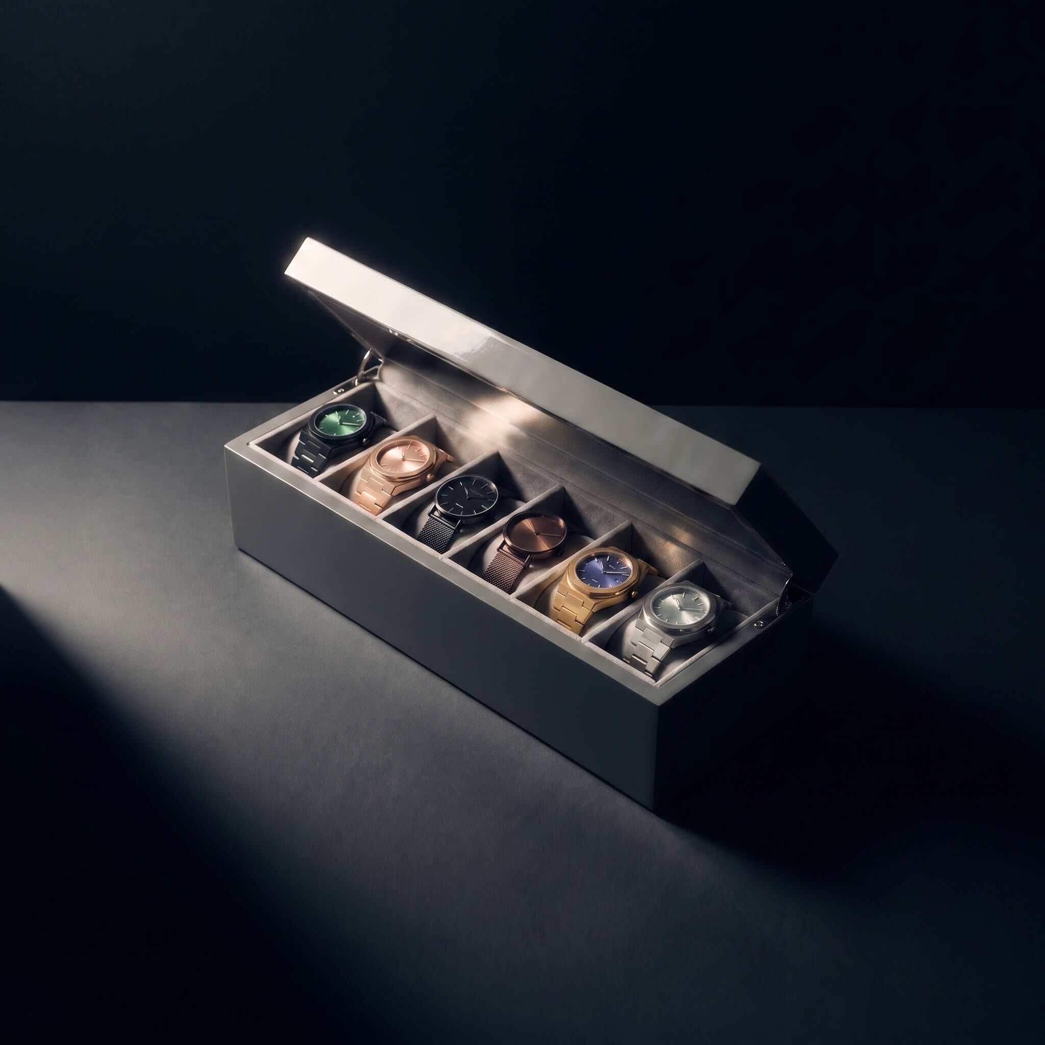 A luxury watch display box with an open lid, showcasing six different wristwatches in individual slots. The box is a dark gray wood with a brushed silver metal lid, set against a dark background with dramatic spotlighting.