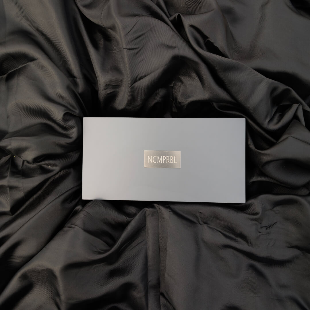 A silver rectangular box with a metallic gold 'NCMPRBL' logo, placed on a dark black satin fabric background.