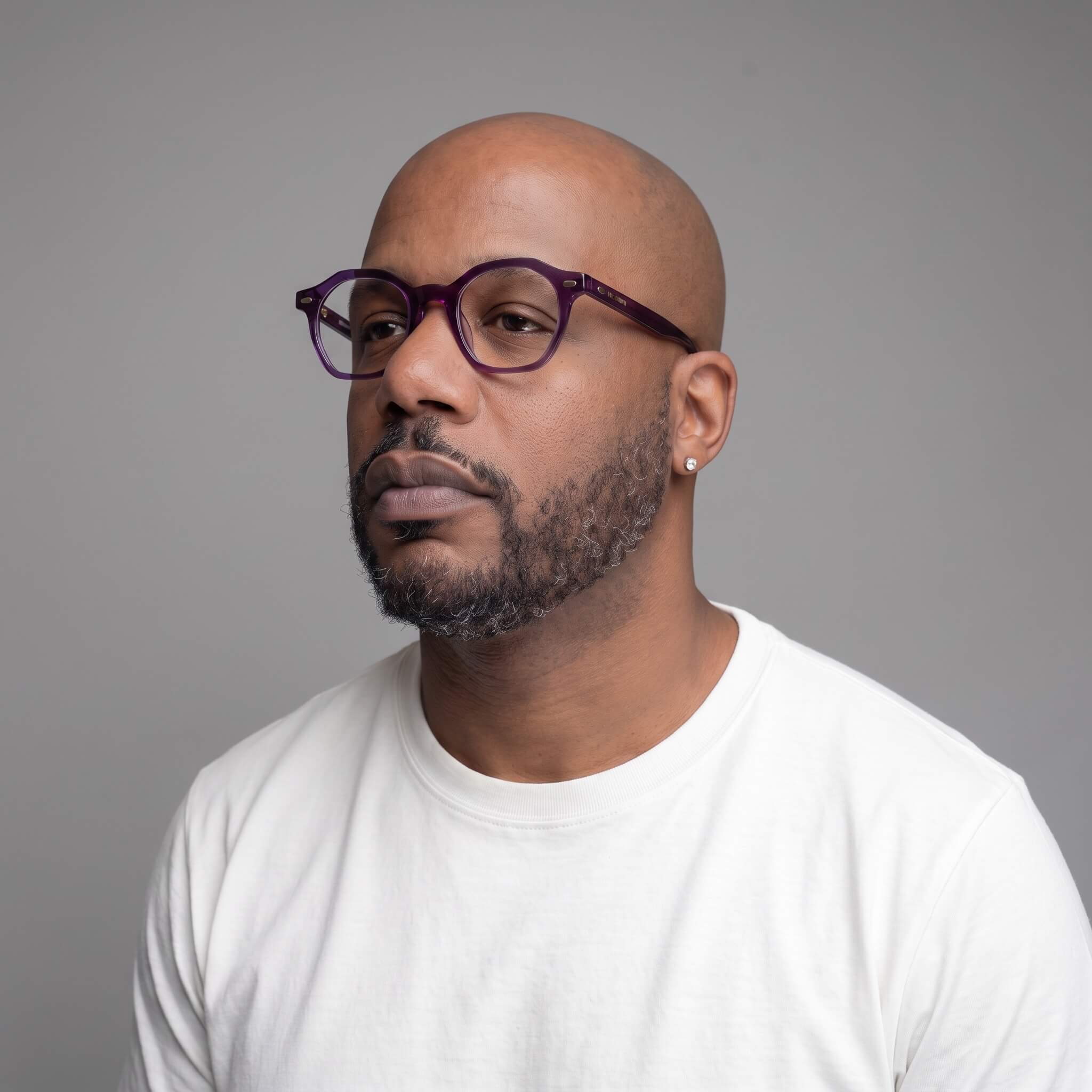 A man with a shaved head and beard wearing haze 46mm purple-framed eyeglasses and a white t-shirt, photographed against a solid gray background.