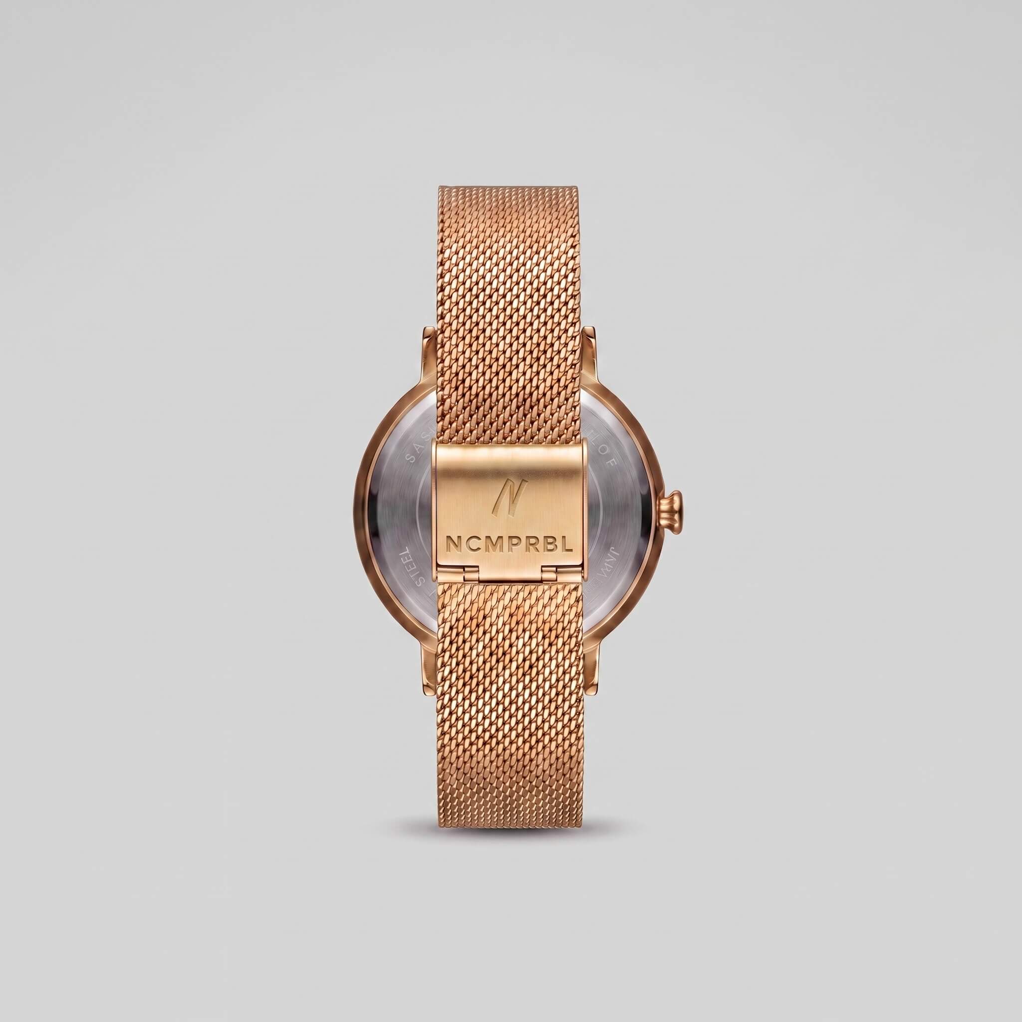 Yellow gold men's watch shown from the back, featuring a round case, mesh strap, and a central metal plate engraved with the brand name 'NCMPRBL'.