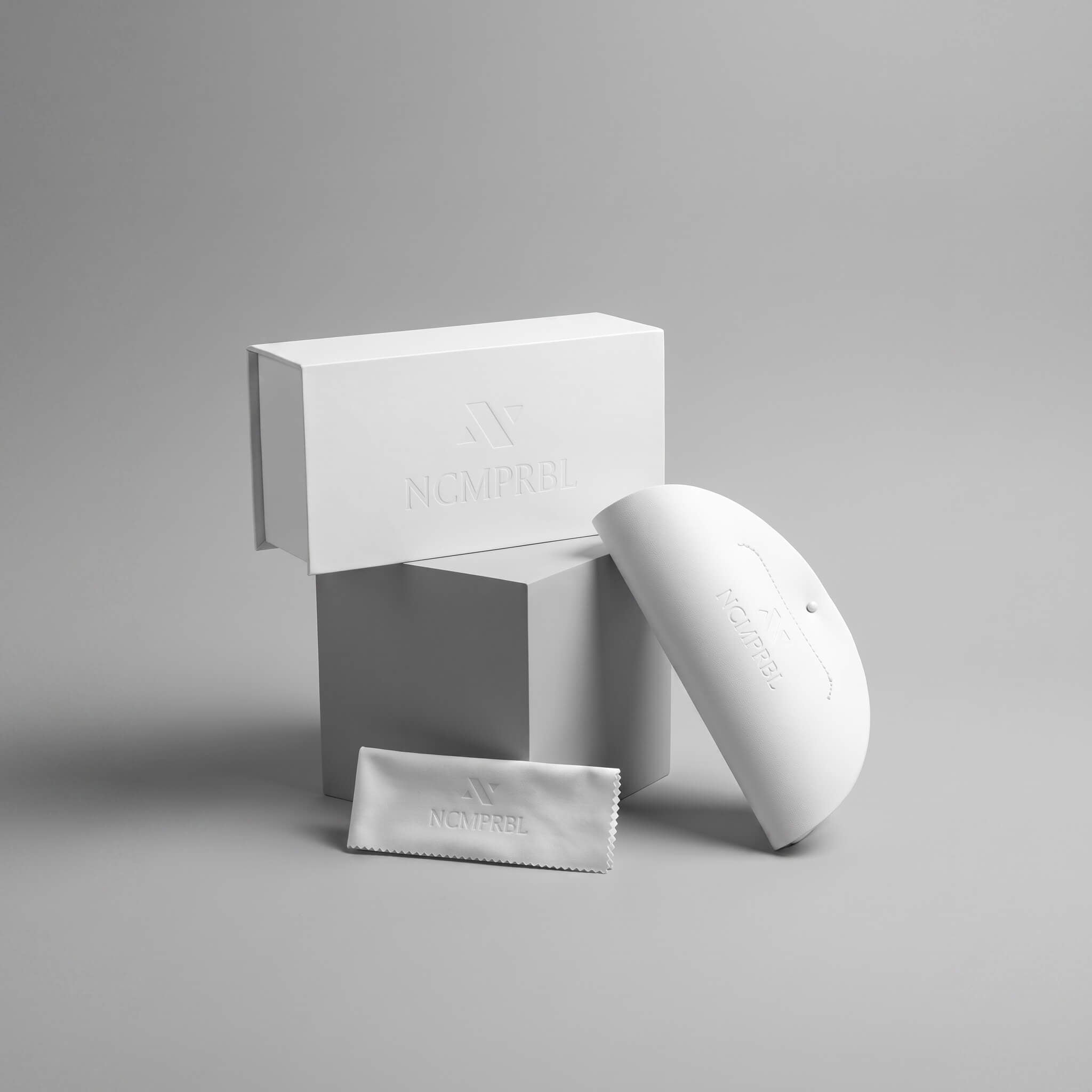A white curved personal care device shown with its matching white box and a small branded pouch on a clean light gray studio background. The embossed branding reads NCMPRBL.