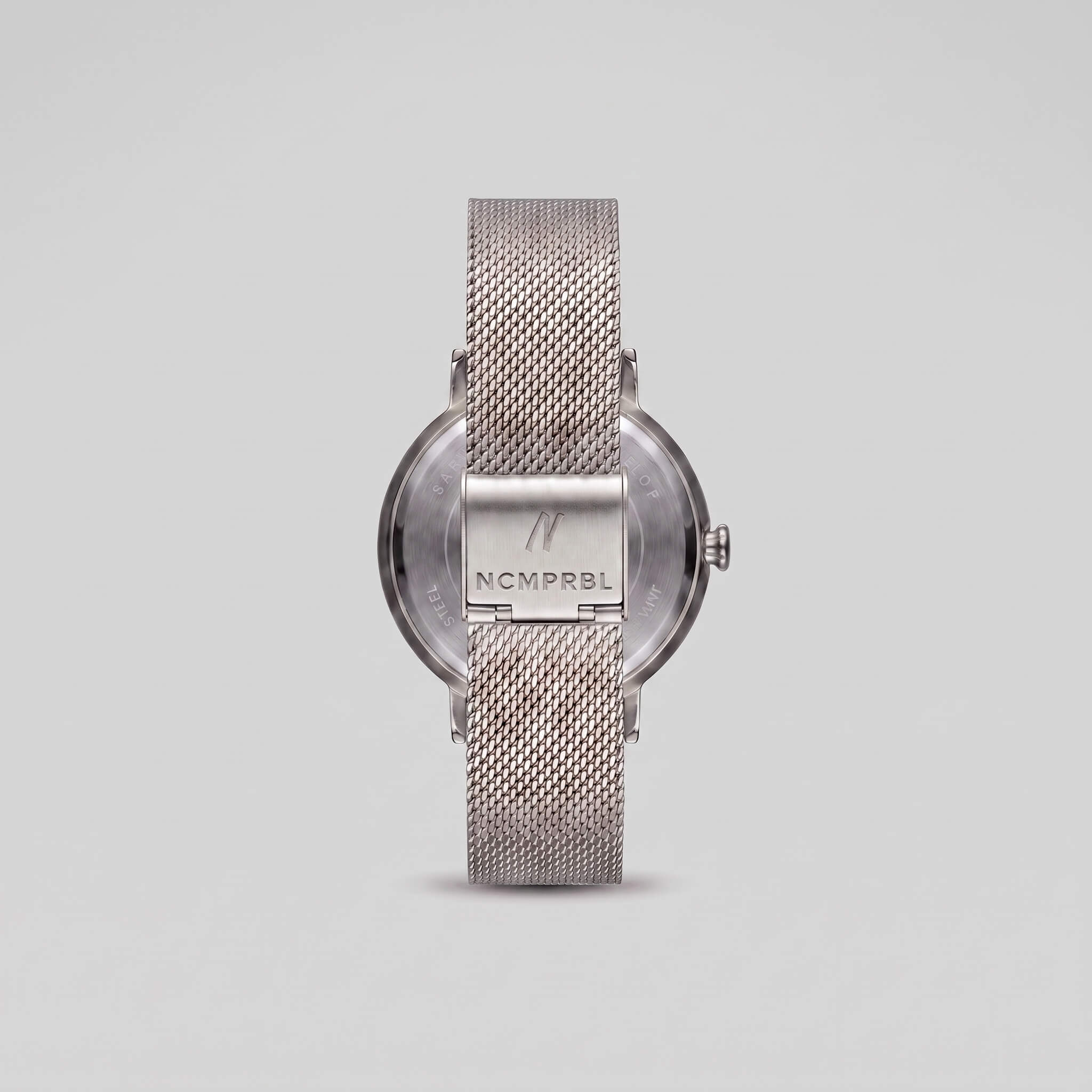 Silver-tone stainless steel watch with a mesh strap, shown from the back on a light gray background. The brand name 'NCMPRBL' is engraved on the center back plate.