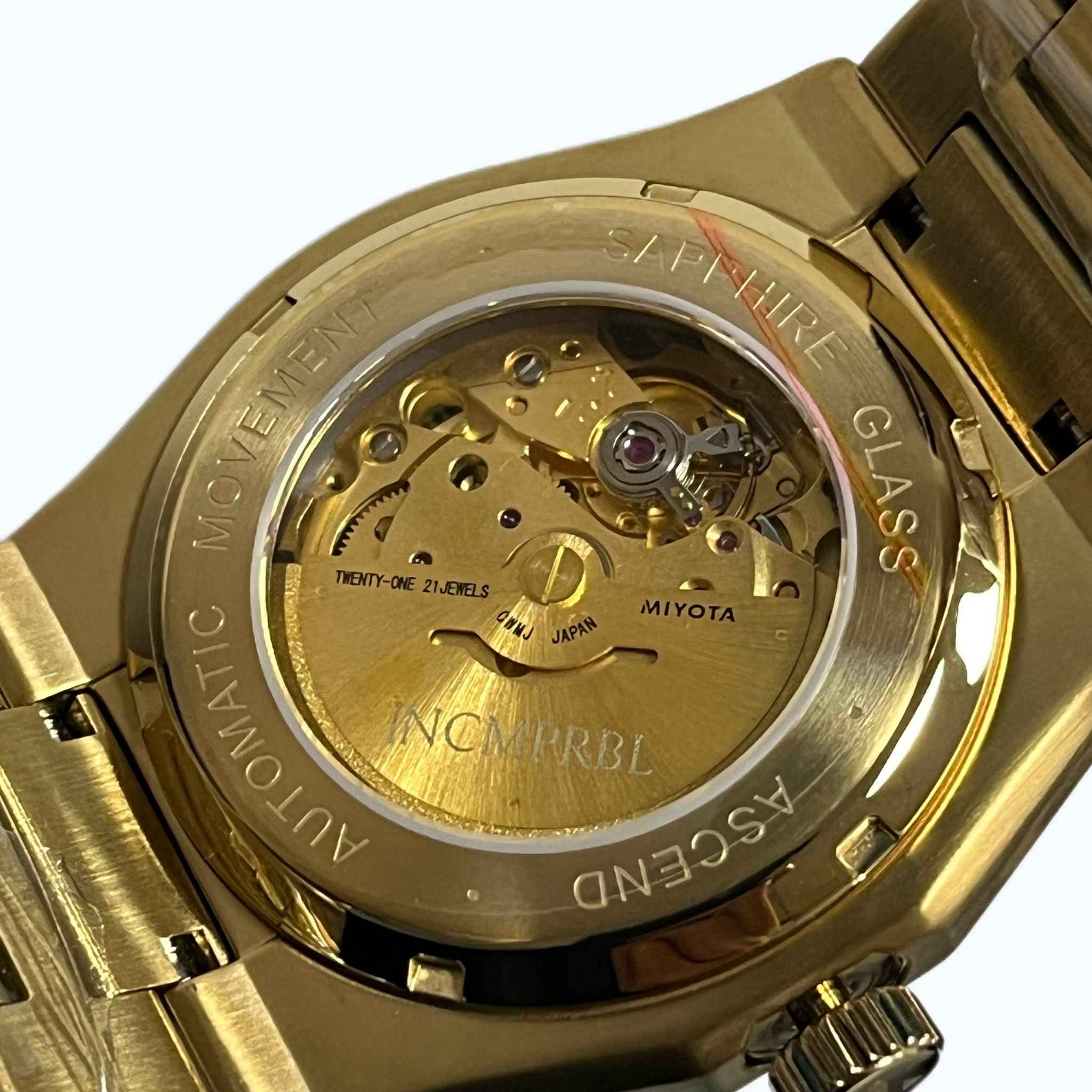 Close-up of the back of a gold-tone wristwatch showing the exposed mechanical movement through a sapphire glass case back. Visible text includes MIYOTA and INCMPRBL.