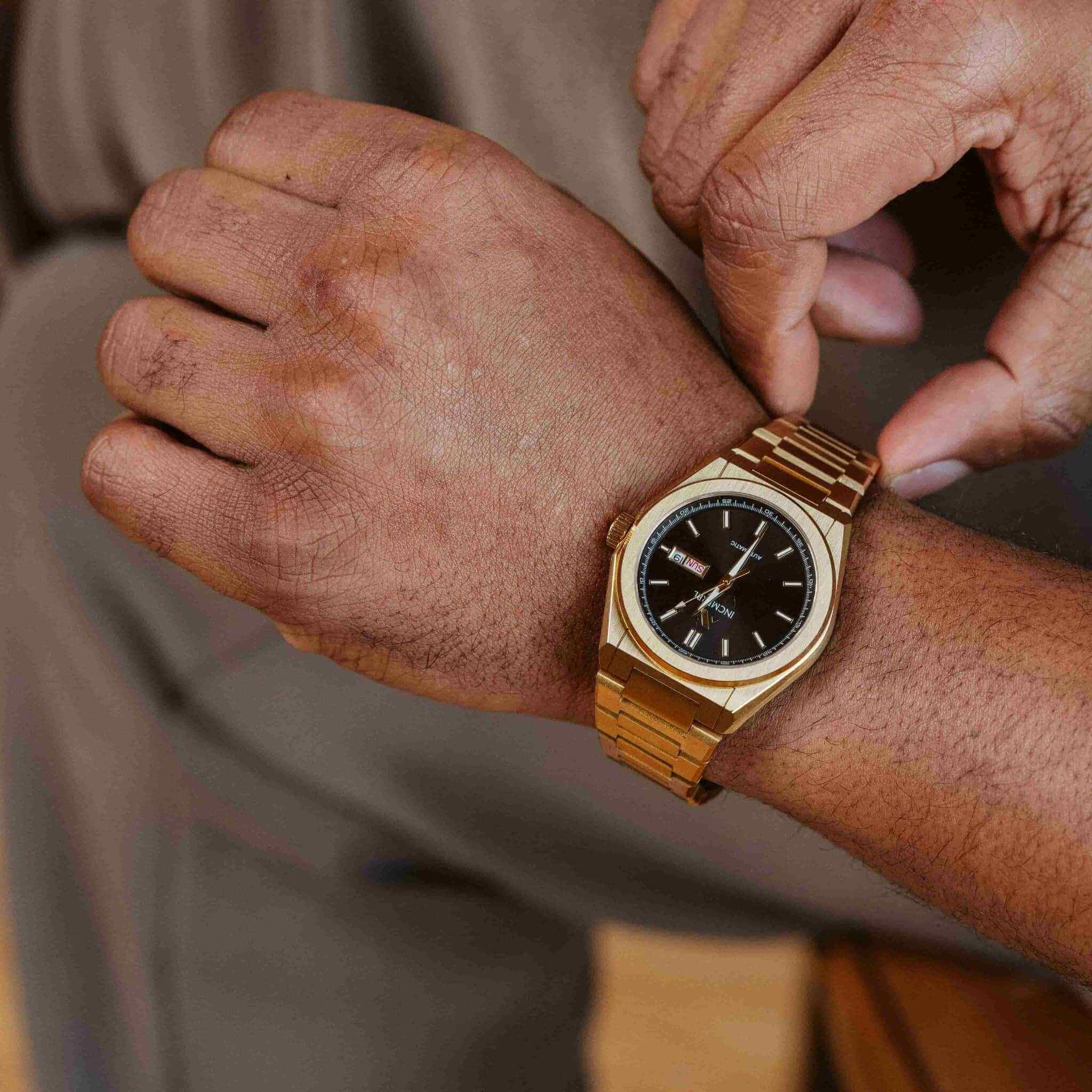 A close-up lifestyle shot of a gold-tone wristwatch with a black dial being worn on a person's wrist, with their other hand adjusting or touching the strap.