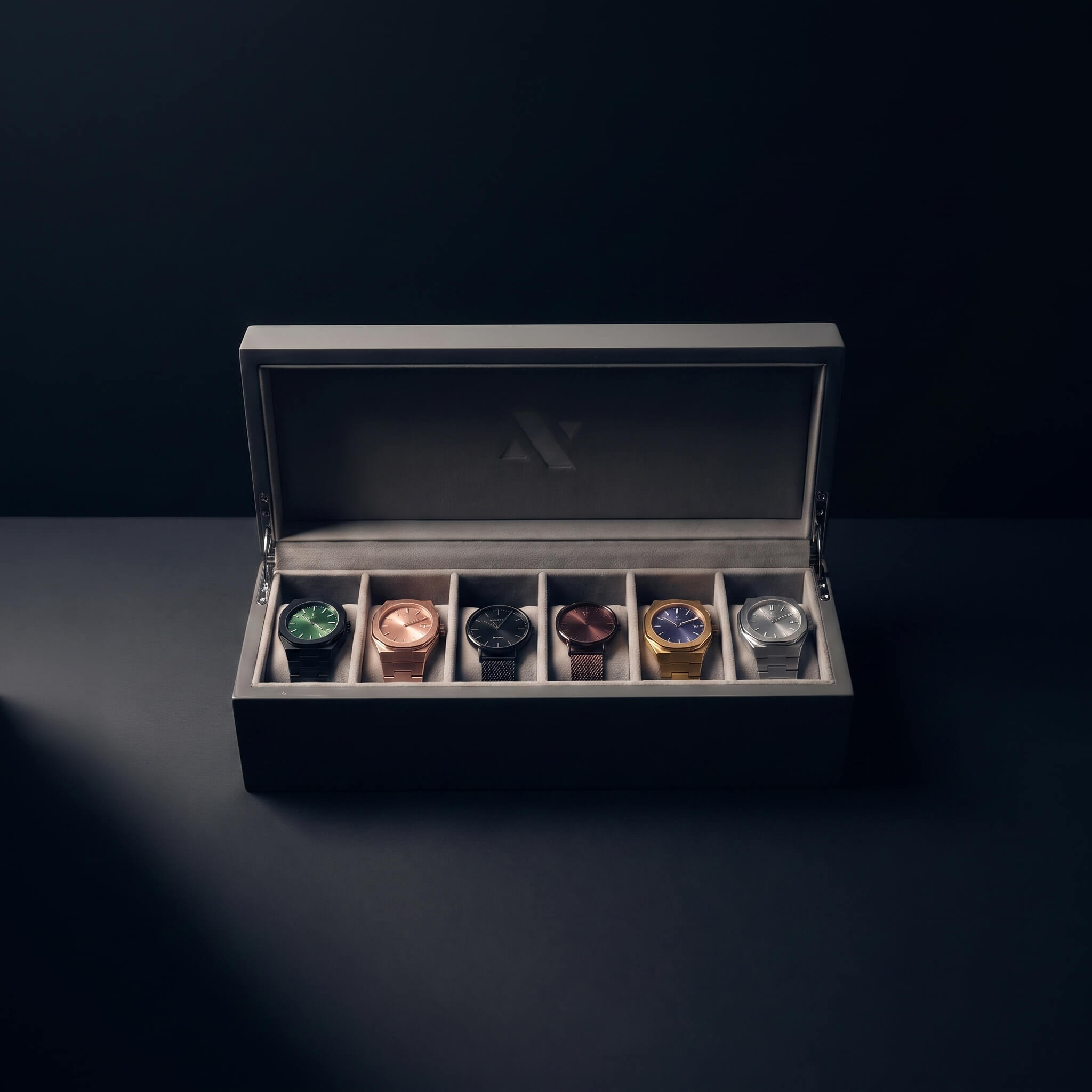 A luxury watch display box with an open lid, showcasing six different wristwatches in individual slots. The watches feature various metal cases (green, rose gold, black, copper, gold, and silver) and dials.