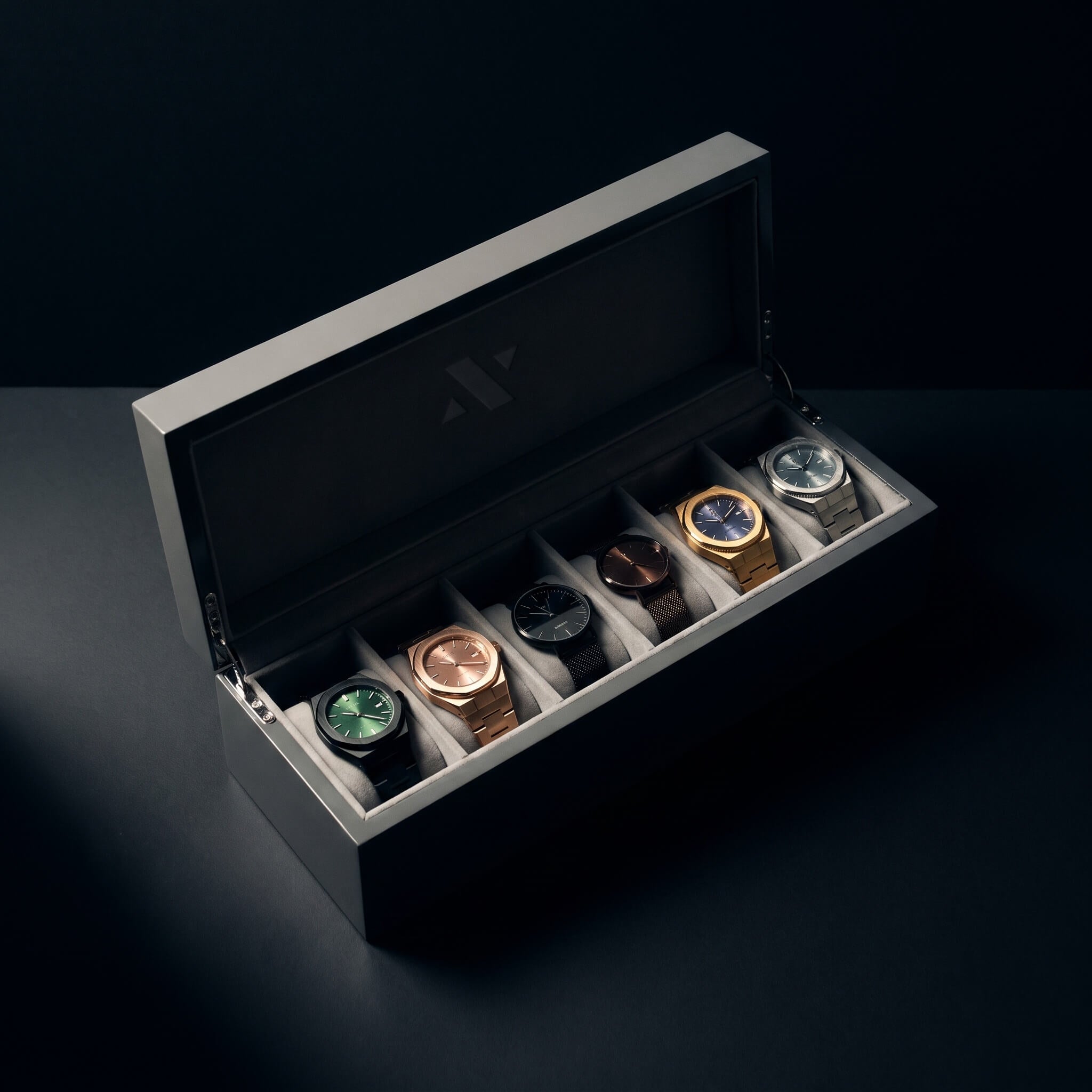 A luxury watch display box with six compartments, shown open to reveal five watches inside in various metal cases (rose gold, yellow gold, silver, black, and green). The box itself is a matte gray with a dark interior, set against a dark, moody background.