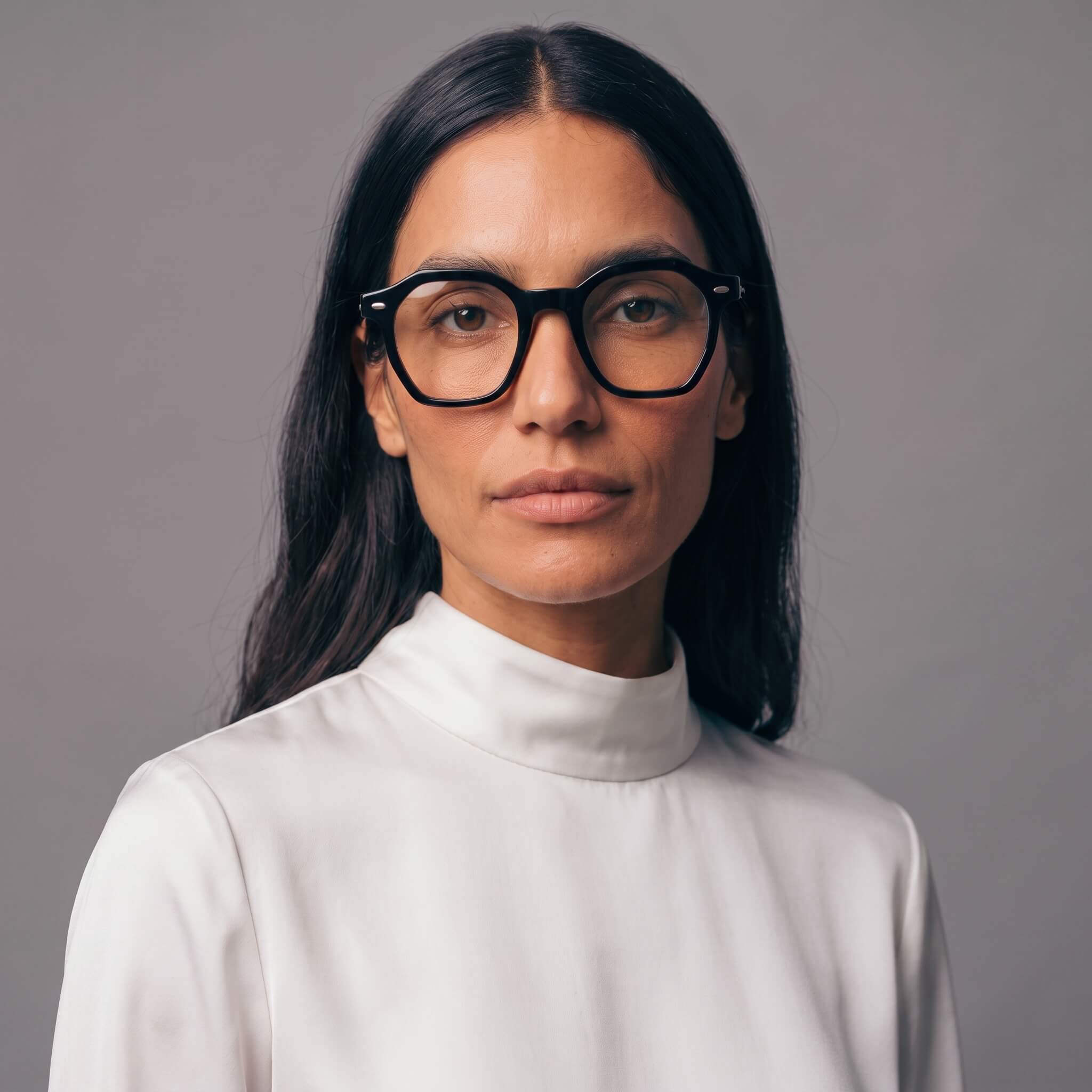 A woman with long dark hair wearing thick black-rimmed eyeglasses and a white mock-neck top, photographed against a plain gray studio background.