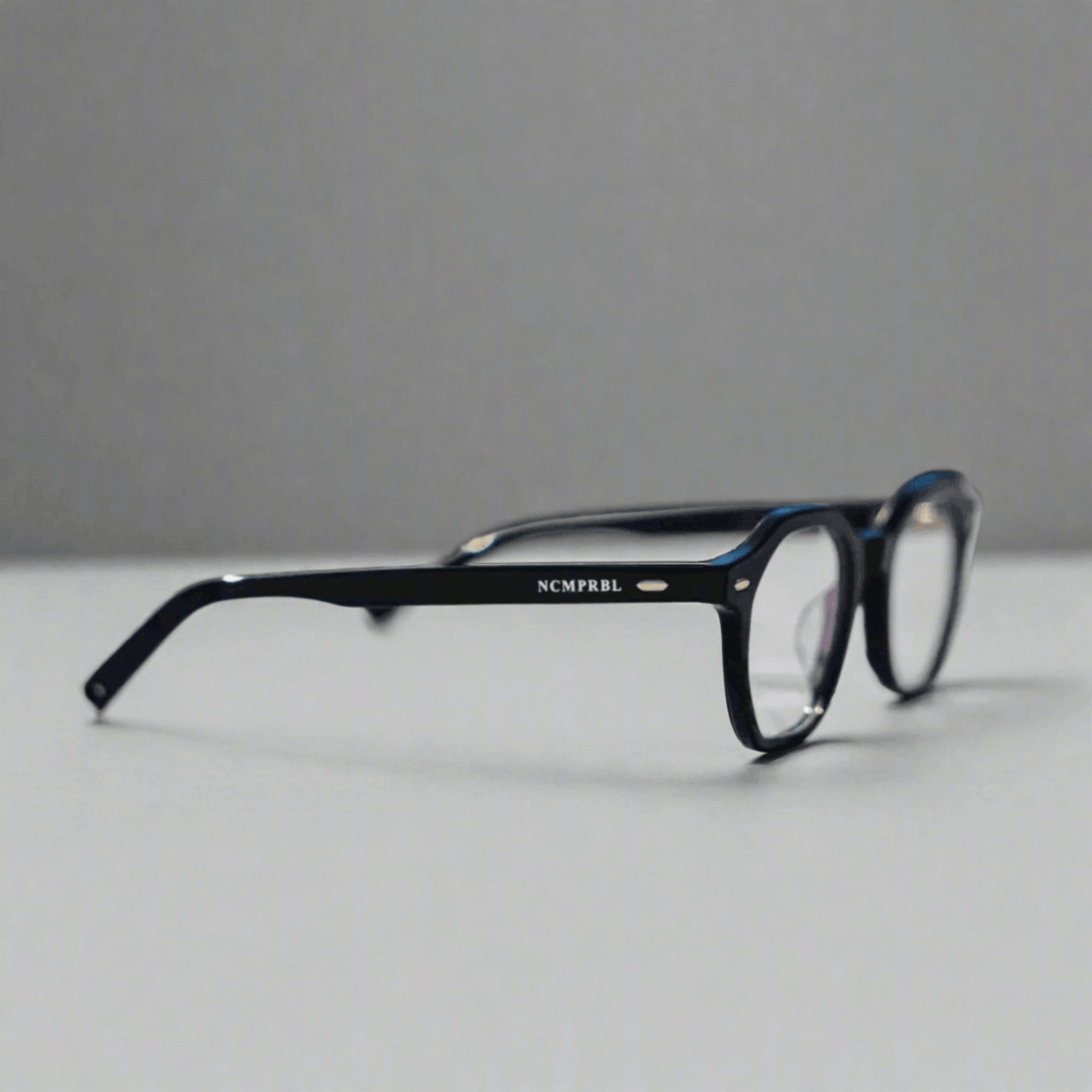 Black acetate eyeglasses with a small silver logo reading 'NCMPRBL' on the temple arm, resting on a light gray surface against a darker gray background.