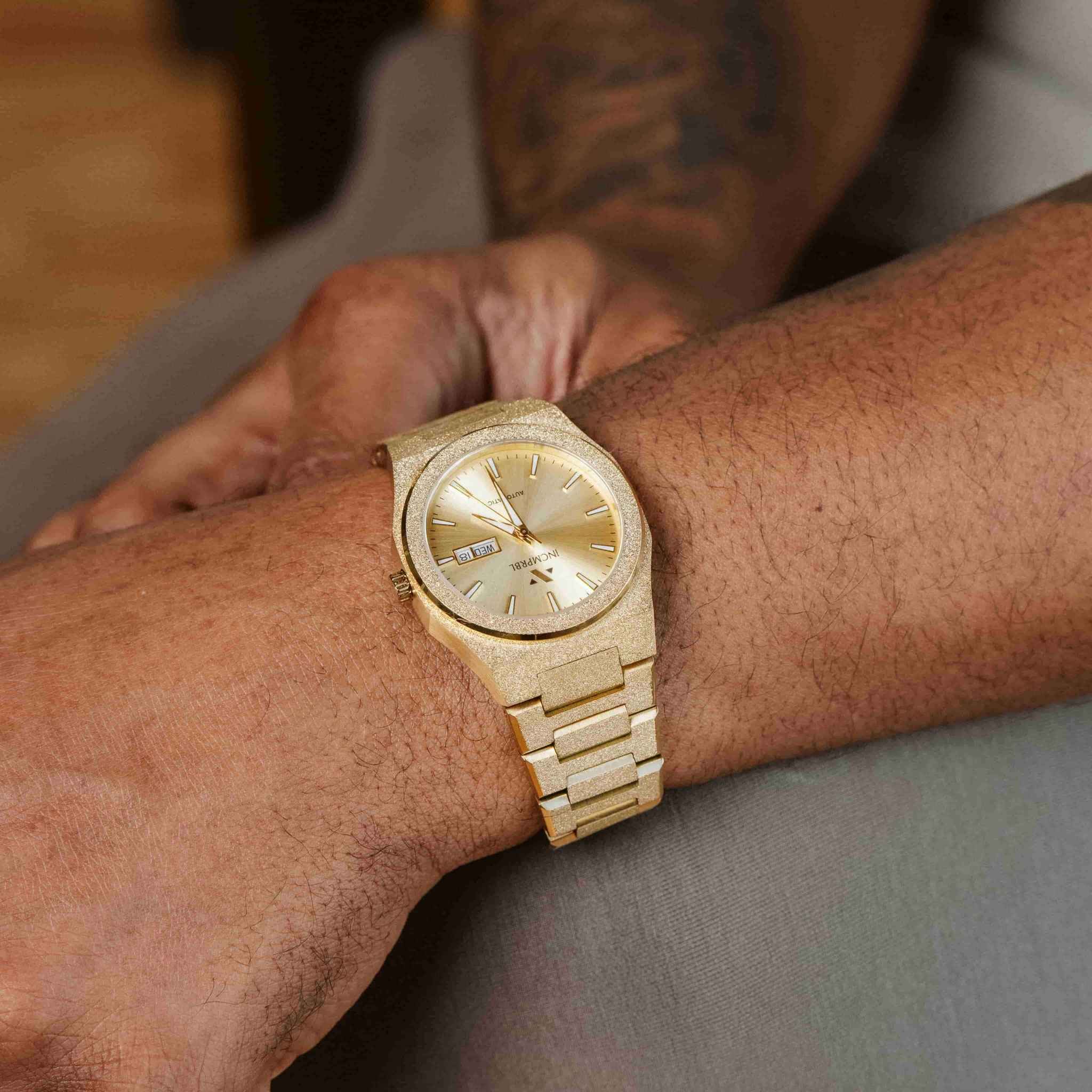 A close-up of a wrist wearing a gold-tone wooden watch with a matching wood bracelet, shown on a person's wrist against a soft gray fabric background.