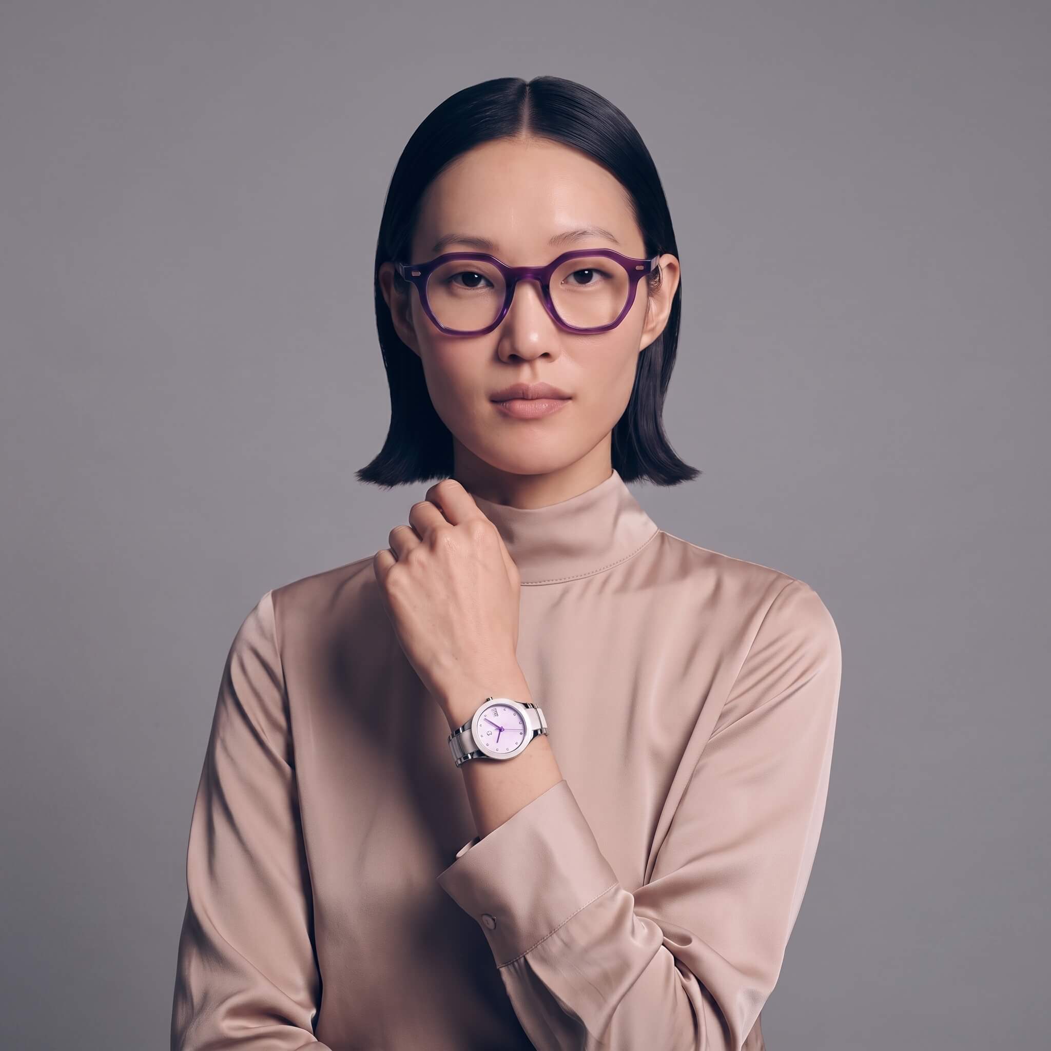 A woman with short dark hair wearing haze 46mm purple-framed eyeglasses, a beige turtleneck top, and a petite link purple reign silver watch with a white dial, posed against a solid gray studio background.