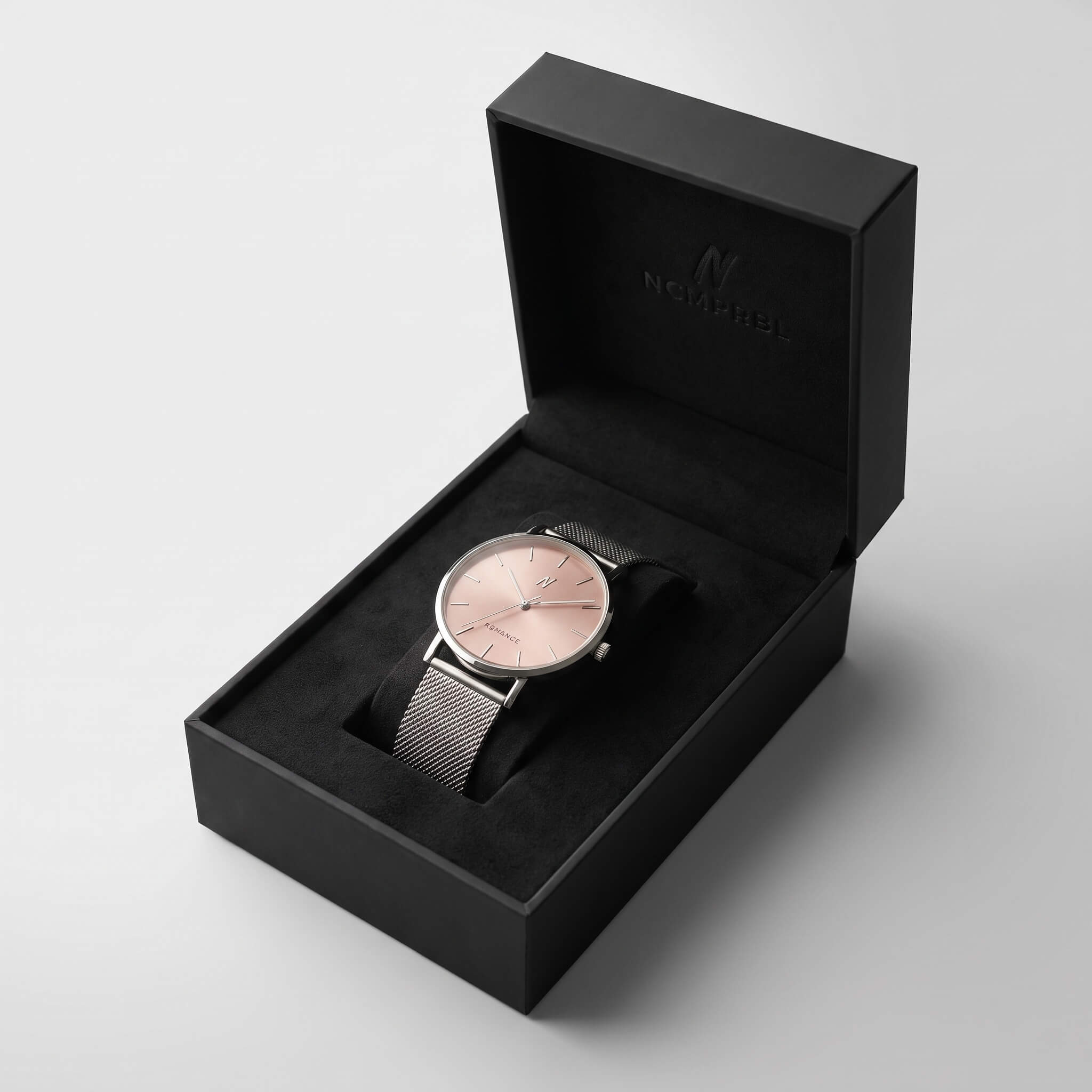A minimalist analog watch with a rose gold dial and a silver mesh strap, presented inside an open black luxury gift box with the brand name 'NCMPRBL' visible on the inner lid.