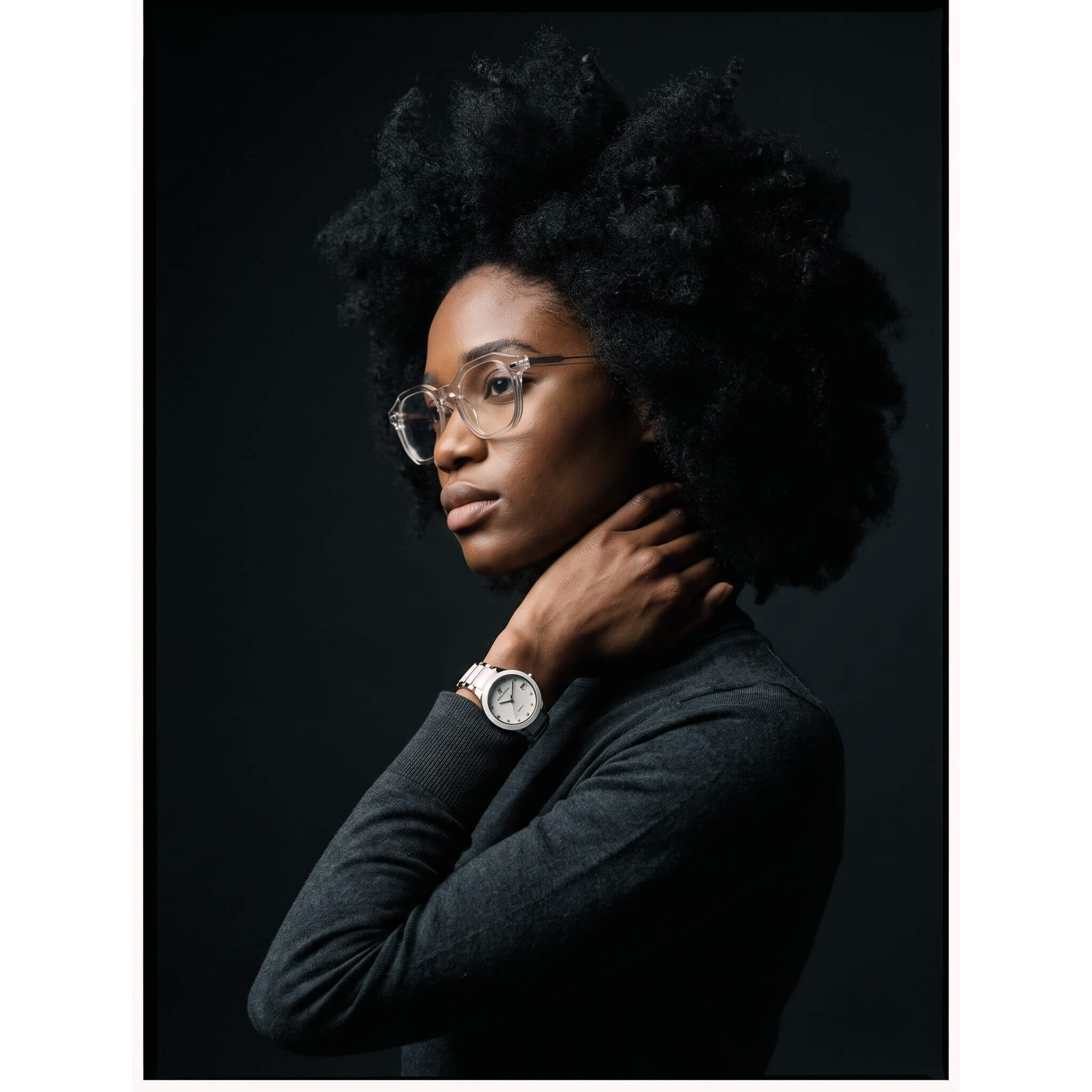 A woman with an afro hairstyle, wearing round eyeglasses, a dark gray long-sleeve top, and a silver watch with a white face, posed against a dark studio background.