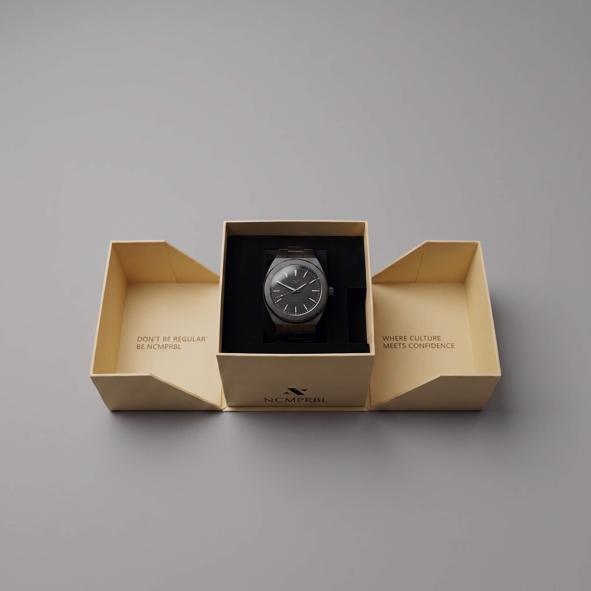 A elevate link phantom wristwatch presented in an open cream-colored branded box on a smooth gray background. The watch has a dark dial and a dark strap, centered inside a black insert, with the brand name NCMPRBL printed on the front of the box.