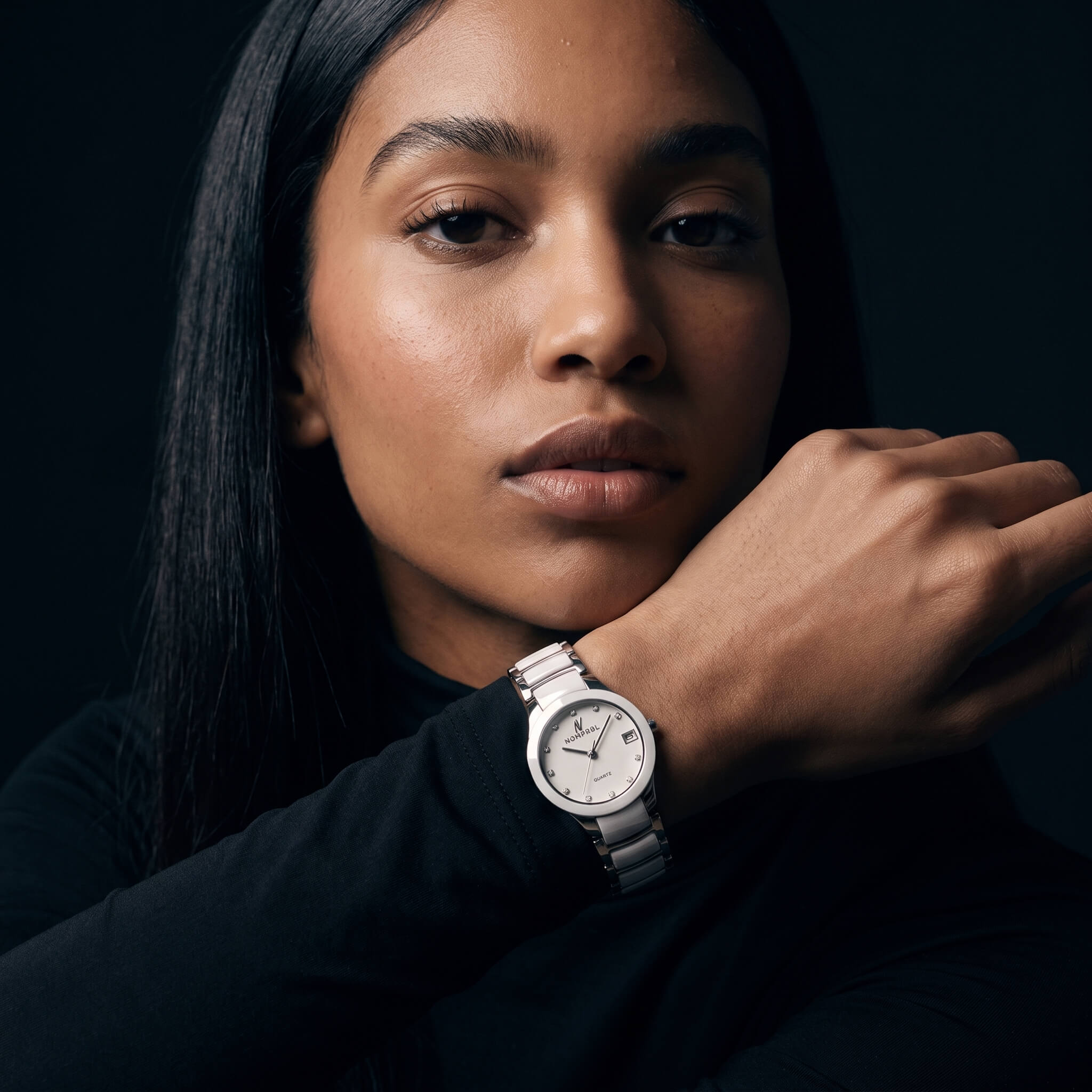 A close-up portrait of a woman wearing a silver-toned stainless steel watch with a white dial and silver link bracelet, set against a dark, moody background.