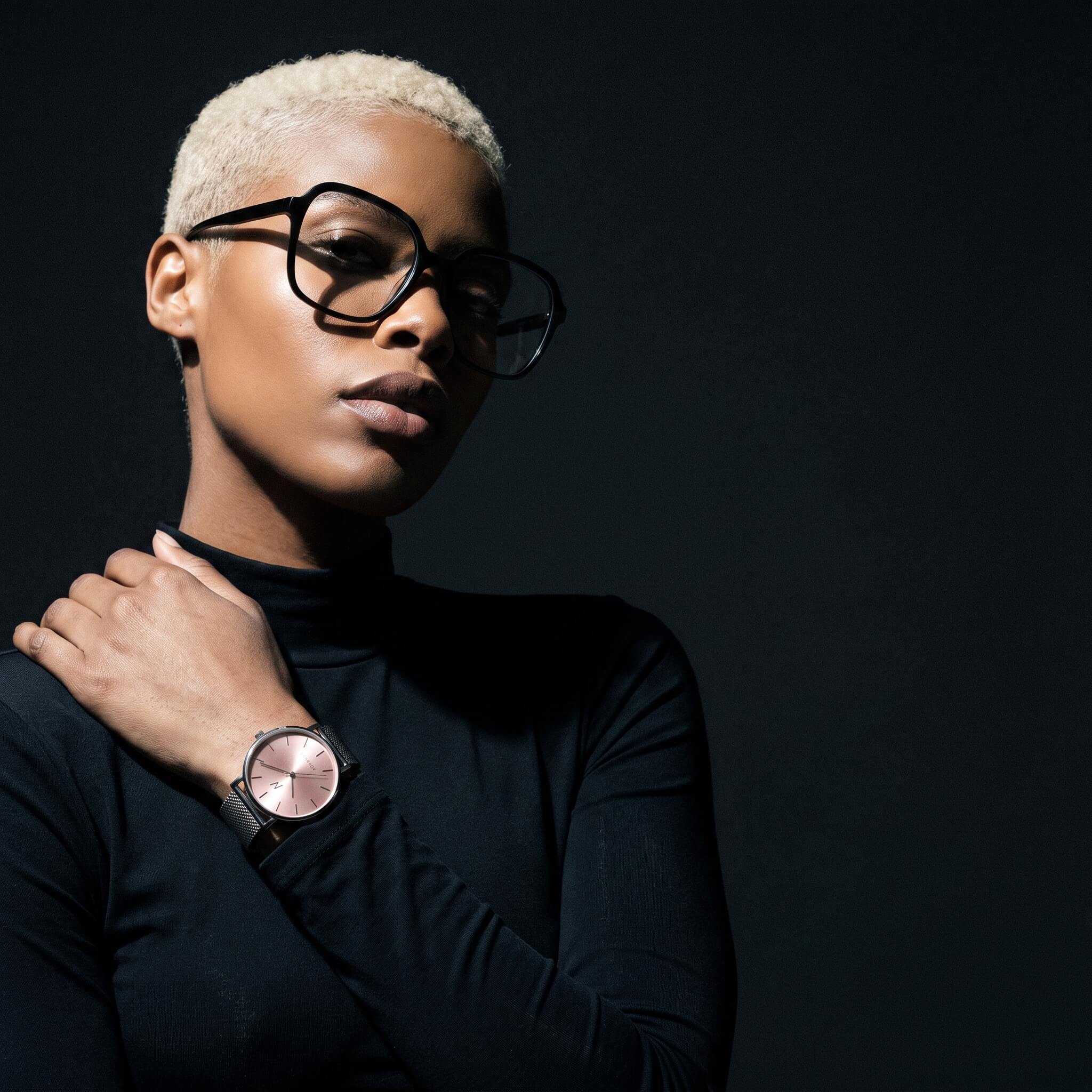 A model with short blonde hair and black-rimmed glasses wearing a Advance Rose Clay Quartz 36MM luxury womens mesh watch with silver pink minimalist design with silver mesh strap, posed against a dark studio background.
